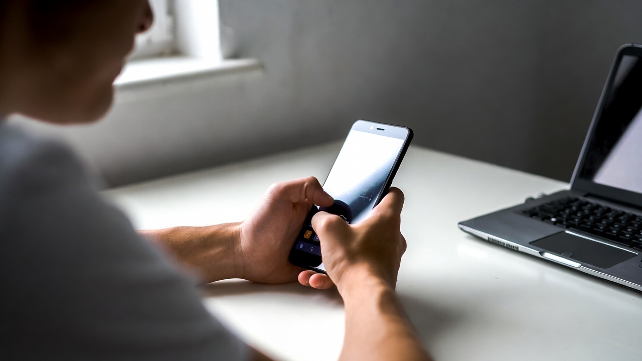 A person sitting at a clean desk using a Xiaomi smartphone with both hands — screen engaged, natural window light from the left side, closed laptop visible in the soft background