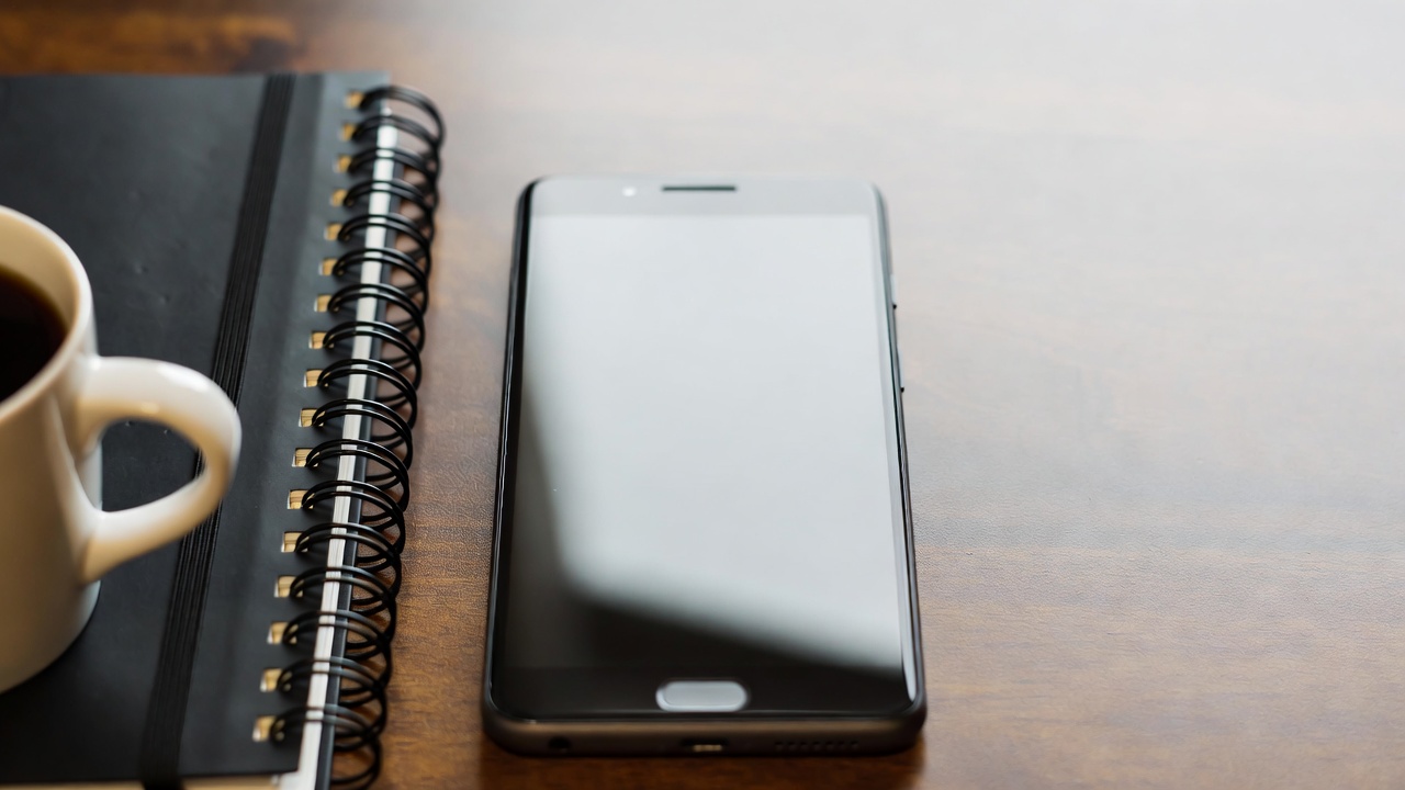 Overhead flat-lay of a smartphone placed on a wooden desk next to a notebook, pen, and a cup of coffee